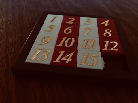 A sliding puzzle consisting of numbered tiles arranged in a 4x4 grid with one missing tile. The tiles are alternately colored red and white, and are encased in a dark brown frame. The numbers are embossed in a gold color.