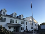 A white two-story building with a gabled roof and multiple windows is accompanied by a tall flagpole displaying an American flag. The building features an entrance with columns and is surrounded by some outdoor chairs and minimal landscaping. The sky is clear and blue, suggesting a bright and sunny day.