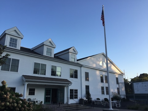 A white two-story building with a gabled roof and multiple windows is accompanied by a tall flagpole displaying an American flag. The building features an entrance with columns and is surrounded by some outdoor chairs and minimal landscaping. The sky is clear and blue, suggesting a bright and sunny day.