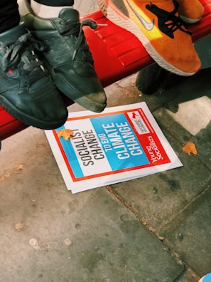 A close-up view of three feet wearing sneakers, two black and one orange, resting on a red bench. Below the bench is a pamphlet with the text 'SOCIALIST CHANGE TO END CLIMATE CHANGE', along with autumn leaves scattered on a sidewalk.