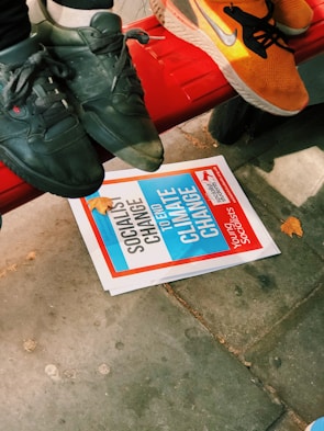 A close-up view of three feet wearing sneakers, two black and one orange, resting on a red bench. Below the bench is a pamphlet with the text 'SOCIALIST CHANGE TO END CLIMATE CHANGE', along with autumn leaves scattered on a sidewalk.