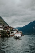 A picturesque lakeside village with charming houses built on a hillside, surrounded by lush green mountains. A white boat is cruising on the calm waters of the lake under an overcast sky filled with grey clouds.