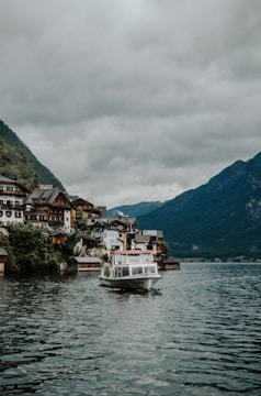 A picturesque lakeside village with charming houses built on a hillside, surrounded by lush green mountains. A white boat is cruising on the calm waters of the lake under an overcast sky filled with grey clouds.
