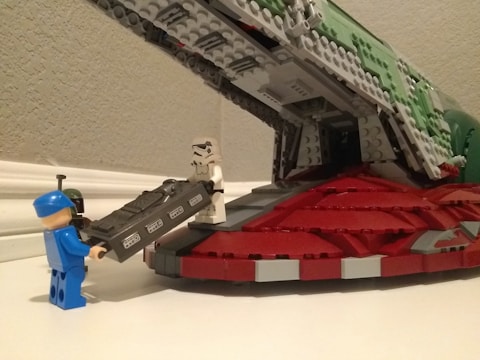 Several LEGO figures are engaged in a scene involving a spacecraft. A figure dressed in a white and black suit resembling a stormtrooper is standing at the ramp of a red and green LEGO ship, which appears detailed and structured. Another figure, wearing a blue outfit and hat, is assisting with carrying a LEGO block that resembles a carbonite slab.