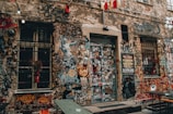 A heavily graffiti-covered urban wall featuring numerous colorful artistic expressions, stickers, and posters. Two windows with metal bars flanking a door plastered with a variety of stickers. A large poster is visible, advertising an event with a date of 22 April 2019. The scene includes strings of lights hanging above, creating an eclectic and vibrant street art atmosphere.