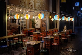 A traditional Japanese street food stall is dimly lit by colorful paper lanterns hanging above. The stall is set with small tables and stools made from repurposed crates, creating an inviting atmosphere despite the dark surroundings. Japanese characters on banners and signs add cultural authenticity to the scene.