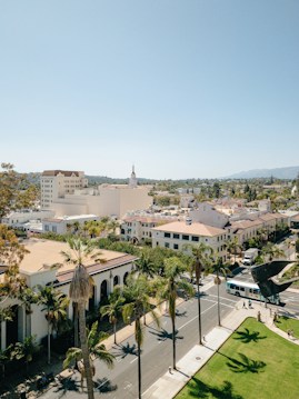 Aerial view of a small town with a mix of historic and modern buildings, palm trees lining the streets, and a clear blue sky. The landscape in the background includes distant mountains.