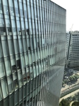 Tall modern building with a glass facade featuring a grid-like pattern. The structure is adjacent to other office buildings and overlooks a cityscape with a mix of greenery and roads.