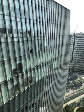 Tall modern building with a glass facade featuring a grid-like pattern. The structure is adjacent to other office buildings and overlooks a cityscape with a mix of greenery and roads.