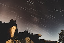 Long exposure photograph capturing the movement of stars across the night sky, creating streaks of light. Silhouetted rocky formations in the foreground add depth and contrast to the celestial scene.