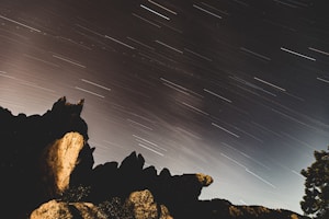 Long exposure photograph capturing the movement of stars across the night sky, creating streaks of light. Silhouetted rocky formations in the foreground add depth and contrast to the celestial scene.