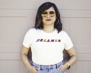 A person stands confidently with hands in pockets, wearing a white T-shirt with the word 'DREAMER' written in colorful letters. They are also wearing jeans, large hoop earrings, sunglasses, and a smartwatch, standing against a muted beige background.