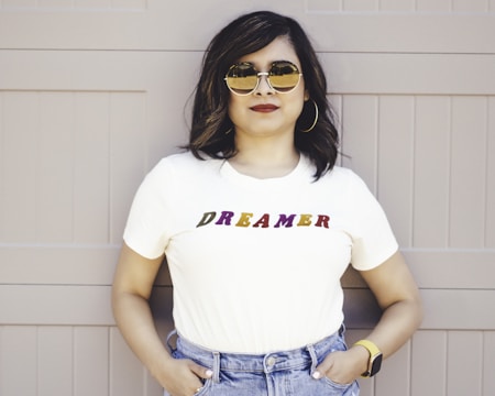 A person stands confidently with hands in pockets, wearing a white T-shirt with the word 'DREAMER' written in colorful letters. They are also wearing jeans, large hoop earrings, sunglasses, and a smartwatch, standing against a muted beige background.