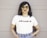 A person stands confidently with hands in pockets, wearing a white T-shirt with the word 'DREAMER' written in colorful letters. They are also wearing jeans, large hoop earrings, sunglasses, and a smartwatch, standing against a muted beige background.