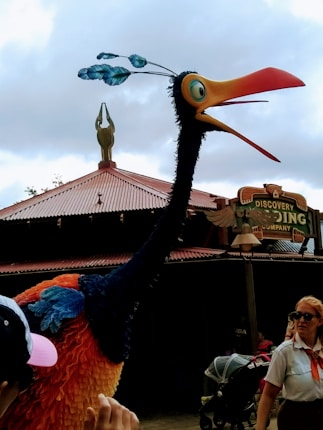 A large, colorful bird-like creature with a long neck and brightly colored plumage, including orange and blue feathers, stands in front of a building with a corrugated roof. A few people are nearby, including a person in uniform and another wearing a cap. A sign in the background reads 'Discovery.' The sky is overcast, creating a muted ambiance.