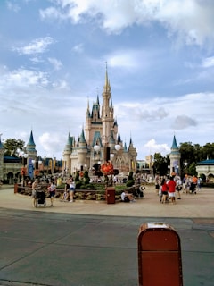 A majestic fairytale castle with multiple spires and turrets stands in the center of a theme park. The area is bustling with visitors, some walking and others gathered in groups, surrounded by decorative gardens and banners. The sky is partially cloudy, providing a picturesque backdrop.