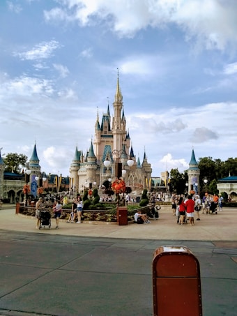 A majestic fairytale castle with multiple spires and turrets stands in the center of a theme park. The area is bustling with visitors, some walking and others gathered in groups, surrounded by decorative gardens and banners. The sky is partially cloudy, providing a picturesque backdrop.