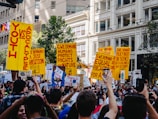 A large group of people are gathered in an urban setting, holding bright yellow protest signs with bold red text. The surroundings include tall buildings and greenery. The protest signs carry various messages demanding action on issues related to climate change and equality. The crowd displays a sense of unity and urgency.
