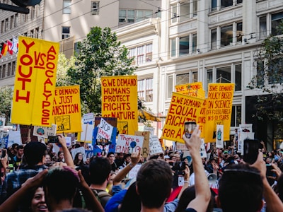 A large group of people are gathered in an urban setting, holding bright yellow protest signs with bold red text. The surroundings include tall buildings and greenery. The protest signs carry various messages demanding action on issues related to climate change and equality. The crowd displays a sense of unity and urgency.