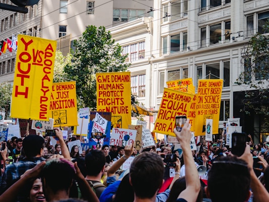 A large group of people are gathered in an urban setting, holding bright yellow protest signs with bold red text. The surroundings include tall buildings and greenery. The protest signs carry various messages demanding action on issues related to climate change and equality. The crowd displays a sense of unity and urgency.