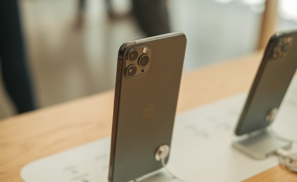 A close-up of two modern smartphones displayed on a wooden surface in an electronics store. Both are tethered with security cables. The focus is on the rear side of the phones, highlighting the camera lenses.