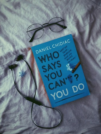 A book with a blue cover titled 'Who Says You Can't? You Do' by Daniel Chidiac is placed on a light-colored fabric surface. Above it are a pair of black-rimmed eyeglasses and next to it is a black wired earphone. A small blue flower decorates the book cover.