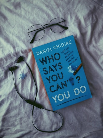 A book with a blue cover titled 'Who Says You Can't? You Do' by Daniel Chidiac is placed on a light-colored fabric surface. Above it are a pair of black-rimmed eyeglasses and next to it is a black wired earphone. A small blue flower decorates the book cover.