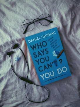 A book with a blue cover titled 'Who Says You Can't? You Do' by Daniel Chidiac is placed on a light-colored fabric surface. Above it are a pair of black-rimmed eyeglasses and next to it is a black wired earphone. A small blue flower decorates the book cover.