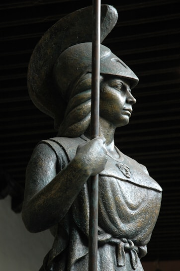 A bronze statue depicts a warrior clad in armor and a helmet, holding a long spear. The figure is shown in a confident pose with detailed craftsmanship visible on the armor and helmet. The background is dark, which contrasts with the metallic textures of the statue.