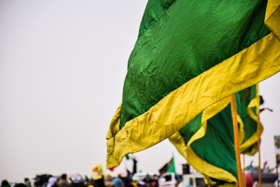 A large green flag with a yellow border is prominently displayed in the foreground. Several similar flags appear in the background, along with a blurred crowd of people and a few vehicles. The sky is overcast, contributing to a muted atmosphere.