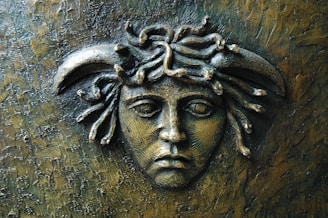 A textured metal relief sculpture features a detailed face with serpentine figures forming the hair, reminiscent of mythological imagery. The expression appears somber, set against an intricately patterned background.