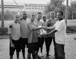 Seven individuals are gathered outdoors on a paved area. They appear to be part of a sports team, wearing casual t-shirts and shorts. Two of them are shaking hands, and one is holding a package. The setting includes palm trees and a building in the background.
