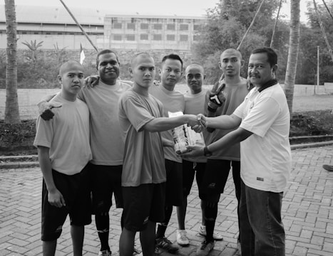 Seven individuals are gathered outdoors on a paved area. They appear to be part of a sports team, wearing casual t-shirts and shorts. Two of them are shaking hands, and one is holding a package. The setting includes palm trees and a building in the background.