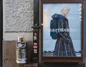 A street scene with a large advertisement featuring a person in a stylish, checked coat carrying a backpack. The ad is mounted on a textured outdoor wall next to a trash bin with several drink cups on top. The area shows signs of urban environment with stickers and minor graffiti.