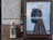A street scene with a large advertisement featuring a person in a stylish, checked coat carrying a backpack. The ad is mounted on a textured outdoor wall next to a trash bin with several drink cups on top. The area shows signs of urban environment with stickers and minor graffiti.