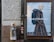 A street scene with a large advertisement featuring a person in a stylish, checked coat carrying a backpack. The ad is mounted on a textured outdoor wall next to a trash bin with several drink cups on top. The area shows signs of urban environment with stickers and minor graffiti.