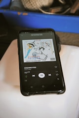 A smartphone is displaying a music player app with the song 'Cigarettes' by Amir Obe playing. The album art is also visible, featuring abstract artwork on a white background. The phone is placed on a light-colored surface, and in the background, there is a blue container with various items that appear to be twine and tools.