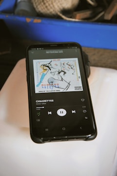 A smartphone is displaying a music player app with the song 'Cigarettes' by Amir Obe playing. The album art is also visible, featuring abstract artwork on a white background. The phone is placed on a light-colored surface, and in the background, there is a blue container with various items that appear to be twine and tools.