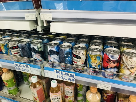 A selection of canned beverages with colorful anime-themed designs on the labels, displayed on a shop shelf. The cans feature various characters and graphics, and some text appears in Japanese. Below the cans are bottles of drinks with visible labels. The price tags in front of the products show prices in what appears to be a yen currency.