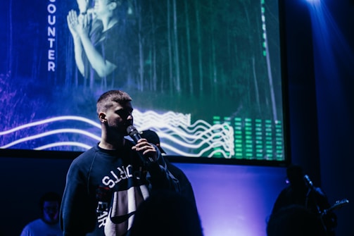 A person holding a microphone stands in front of a large screen displaying abstract visuals, possibly during a presentation or performance. The background features a forest scene with digital waveforms and vertical lines with green highlights, creating a futuristic atmosphere.