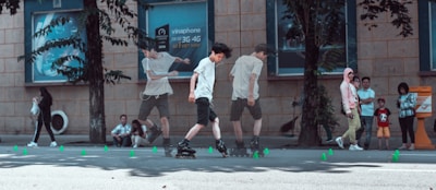 A young person is rollerblading on a city street, performing tricks and weaving through a series of small green cones. In the background, several people are observing, standing near trees and a building that has signs. The street is lined with trees casting shadows on the pavement.