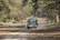 A vehicle with a safari banner drives down a dirt path surrounded by dense forest. The path is covered with dry leaves, suggesting autumn. Sunlight filters through the trees, creating dappled shadows on the ground.