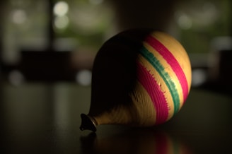 A spinning top is placed on a dark surface with a blurred, out-of-focus background. The top is primarily wooden with vibrant pink, yellow, and green stripes running horizontally.