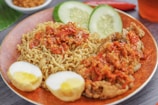 A plate of noodles topped with spicy red chili sauce alongside pieces of fried chicken. There are slices of cucumber and two halves of a boiled egg with a yellow yolk. A small bowl of fried shallots and a red chili pepper are in the background.