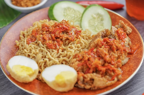 A plate of noodles topped with spicy red chili sauce alongside pieces of fried chicken. There are slices of cucumber and two halves of a boiled egg with a yellow yolk. A small bowl of fried shallots and a red chili pepper are in the background.