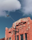 A weathered and peeling facade of a red brick building labeled 'Sandringham Hotel' with exposed plaster and a distressed look. A streetlight rises in front of it, and power lines cross the scene. The sky is clear with a few fluffy white clouds.