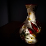 A ceramic vase with a narrow neck and a rounded body features hand-painted floral patterns in red, yellow, and green. The vase is situated in a dark environment, with light casting dramatic shadows on its surface.