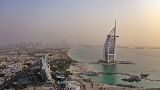 Aerial view of a modern cityscape featuring a prominent sail-shaped building located on an island. Surrounding it are waterways and urban infrastructure, with additional skyscrapers visible in the distance under a hazy sky.