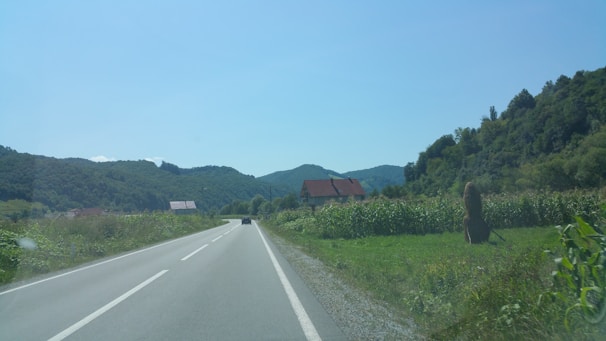 A rural landscape with a winding road flanked by green fields and dense forested hills in the background. A few houses with red roofs are visible alongside the road, and a haystack stands prominently in one of the fields. The sky is clear and blue, adding to the tranquility of the scene.