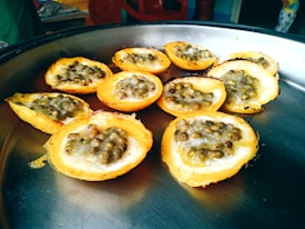 A metal tray holds several round, baked pastries filled with a creamy mixture and topped with small, greenish-brown seeds. These pastries appear slightly browned around the edges, indicating they have been cooked. In the background, a red chair and some household items are slightly visible.
